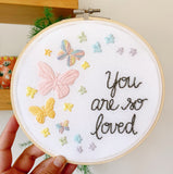 Embroidery hoop being held in my hand that has rainbow pastel butterflies stitched into the fabric with the text, "you are so loved"