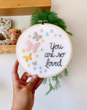 Embroidery hoop being held in my hand that has rainbow pastel butterflies stitched into the fabric with the text, "you are so loved"