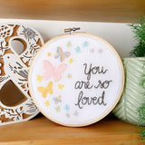 Embroidery hoop on a shelf that has rainbow pastel butterflies stitched into the fabric with the text, "you are so loved"