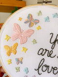 Embroidery hoop on a shelf that has rainbow pastel butterflies stitched into the fabric with the text, "you are so loved"