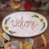 This is an oval embroidery hoop that is stained brown with peach and white floral fabric. The word "welcome" is embroidered on the fabric with yellow flowers and green leaves. The hoop is sitting on a wooden background.