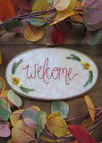 This is an oval embroidery hoop that is stained brown with peach and white floral fabric. The word "welcome" is embroidered on the fabric with yellow flowers and green leaves. The hoop is sitting on a wooden background.