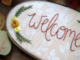 This is an oval embroidery hoop that is stained brown with peach and white floral fabric. The word "welcome" is embroidered on the fabric with yellow flowers and green leaves. The hoop is sitting on a wooden background. 