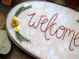 This is an oval embroidery hoop that is stained brown with peach and white floral fabric. The word "welcome" is embroidered on the fabric with yellow flowers and green leaves. The hoop is sitting on a wooden background.