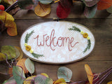 This is an oval embroidery hoop that is stained brown with peach and white floral fabric. The word "welcome" is embroidered on the fabric with yellow flowers and green leaves. The hoop is sitting on a wooden background.