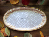 This is an oval embroidery hoop that is stained brown with peach and white floral fabric. The word "welcome" is embroidered on the fabric with yellow flowers and green leaves. The hoop is sitting on a wooden background.