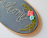 Dark grey fabric in an oval hoop with the word "welcome" embroidered in white. There are pink and blue flowers with green leaves.
