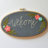Dark grey fabric in an oval hoop with the word "welcome" embroidered in white. There are pink and blue flowers with green leaves.