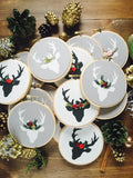 This is a pile of four stag ornaments in black and white with red and green floral crowns. They are on a white wooden surface with pine cones and acorns scattered around in a circle.