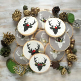 This is a pile of four stag ornaments in black and white with red and green floral crowns. They are on a white wooden surface with pine cones and acorns scattered around in a circle.