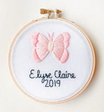 This is a 4 inch embroidery hoop with white fabric and a pink butterfly embroidered in the center. under the butterfly is a baby name stitched in black with the date underneath.