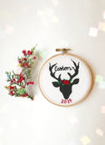 This is a small 4 inch embroidery hoop with a black deer shape printed on white fabric. There is a name embroidered in black, as well as the year in red at the bottom. The deer has a floral crown on its head between the antlers. Besides the deer there is a prop of holly berry.