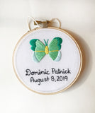 This is a 4 inch embroidery hoop with white fabric and a green and mint butterfly embroidered in the center. under the butterfly is a baby name stitched in black with the date underneath.