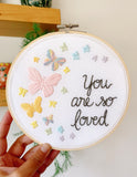 Embroidery hoop being held in my hand that has rainbow pastel butterflies stitched into the fabric with the text, "you are so loved"