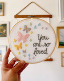 Embroidery hoop being held in my hand that has rainbow pastel butterflies stitched into the fabric with the text, "you are so loved"
