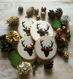 This is a pile of four stag ornaments in black and white with red and green floral crowns. They are on a white wooden surface with pine cones and acorns scattered around in a circle.