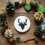 This is a small 4 inch embroidery hoop with a black deer shape printed on white fabric. There is a name embroidered in black, as well as the year in red at the bottom. The deer has a floral crown on its head between the antlers. Around the deer are winter nature props, such as pine cones and cinnamon sticks arranged in a circle.