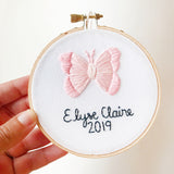 This is a 4 inch embroidery hoop with white fabric and a pink butterfly embroidered in the center. under the butterfly is a baby name stitched in black with the date underneath. 
