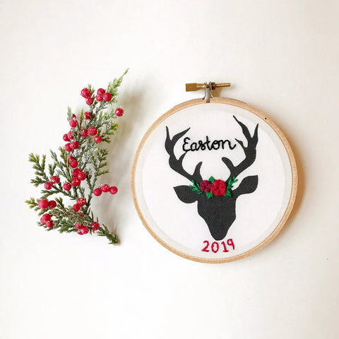 This is a small 4 inch embroidery hoop with a black deer shape printed on white fabric. There is a name embroidered in black, as well as the year in red at the bottom. The deer has a floral crown on its head between the antlers. Besides the deer there is a prop of holly berry.