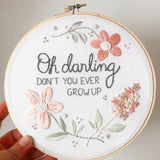 A hand holding a 7 inch embroidery hoop with a white background and flowers in shades of pink and taupe. There is text embroidered in grey that says "oh darling don't you ever grow up".