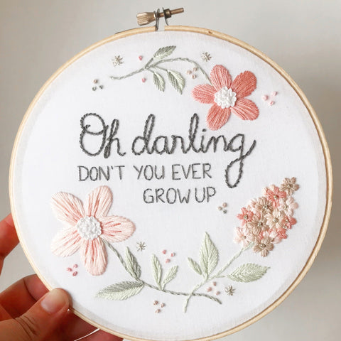 A hand holding a 7 inch embroidery hoop with a white background and flowers in shades of pink and taupe. There is text embroidered in grey that says "oh darling don't you ever grow up".