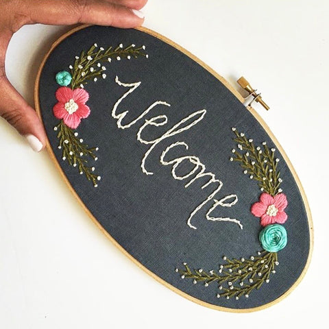 Dark grey fabric in an oval hoop with the word "welcome" embroidered in white. There are pink and blue flowers with green leaves.