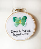 This is a 4 inch embroidery hoop with white fabric and a green and mint butterfly embroidered in the center. under the butterfly is a baby name stitched in black with the date underneath. 